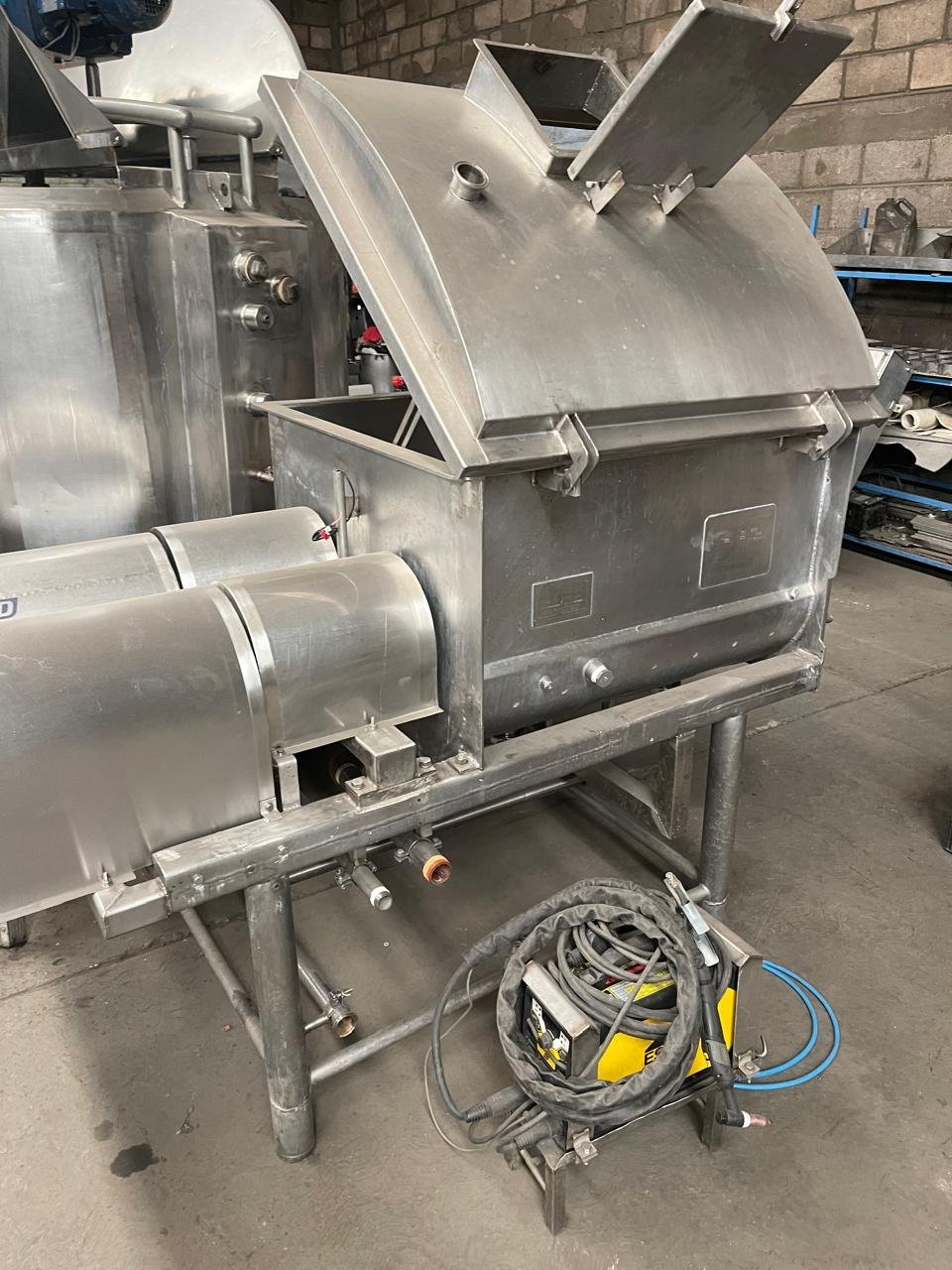 Cooker industrial de acero inoxidable con tapa superior tipo techo, tolva de alimentación y cables enrollados en piso de taller